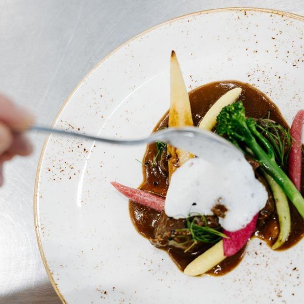 „Hände setzen den letzten Schliff an ein kunstvoll angerichtetes Gourmetgericht mit Sauce, Gemüse und Kräutern auf weißem Teller.